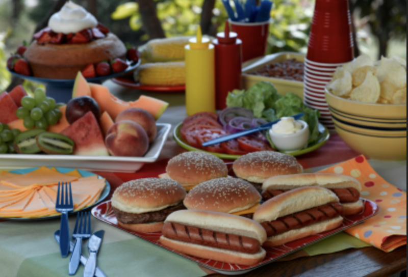 Image of picnic table with hot dogs, hamburgers, condiments and chips